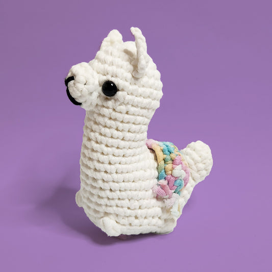 White alpaca crochet amigurumi with a rainbow yarn saddle, made from our beginner-friendly crochet kit. Handcrafted with love, this adorable alpaca is perfect for beginners and alpaca enthusiasts. Front view showcasing its charming design and colorful saddle.