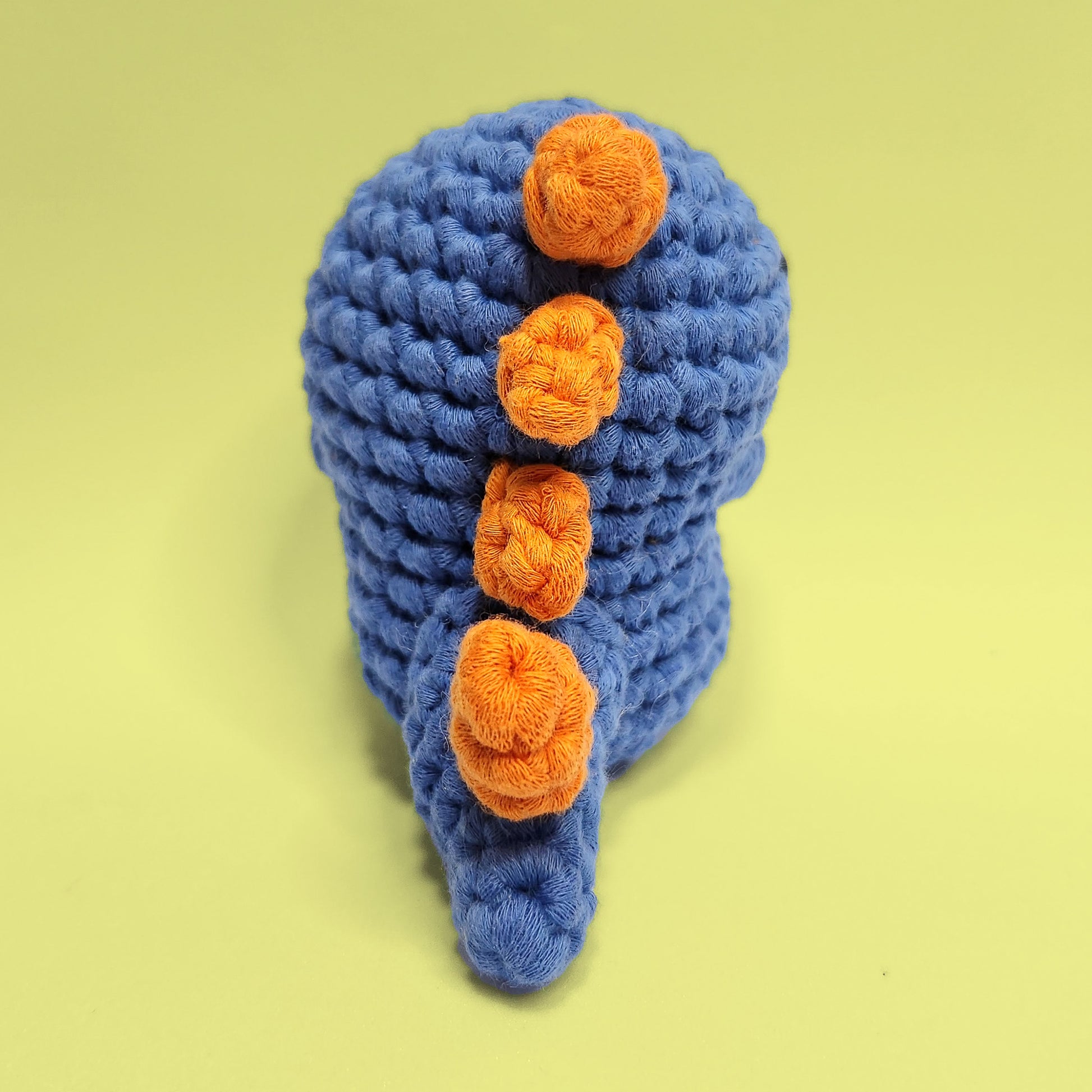 Blue dinosaur crochet amigurumi with a bulging stomach and orange spikes on the back. Handcrafted from our beginner friendly kit, this unique dinosaur is a fun and engaging project for crochet enthusiasts. Back view.